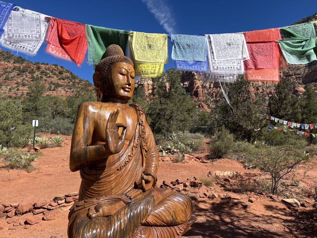 Large wooden Buddha statue seated at an outdoor stupa with trees and red rock landscape